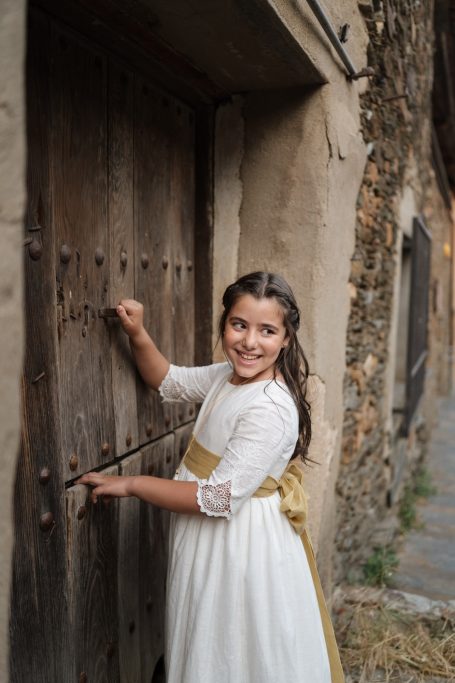 Niña sonriente tocando una puerta antigua en un callejón de piedra.