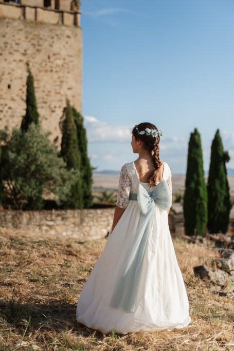 Mujer de espaldas en vestido blanco con lazo azul, frente a un castillo y arbustos.