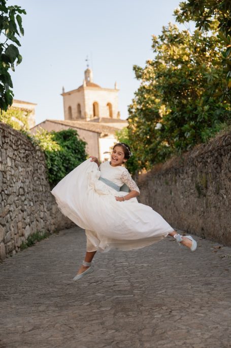 Mujer bailando con vestido blanco en un camino entre árboles y edificios.