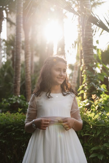 Niña sonriente en un vestido blanco, rodeada de plantas y luz solar brillante.