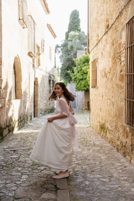 Mujer sonriente con vestido blanco camina por una calle empedrada en un pueblo antiguo.