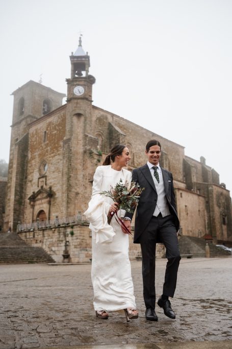 Pareja sonriente caminando juntos, frente a una iglesia antigua en un día nublado.