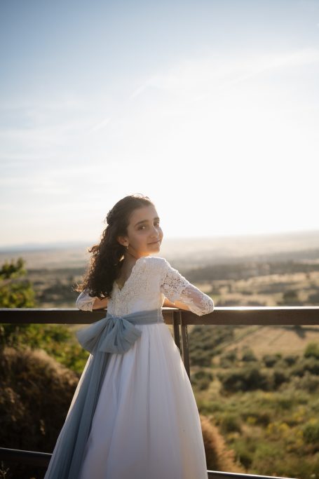 Niña con vestido blanco y lazo azul, mirando hacia el horizonte al atardecer.
