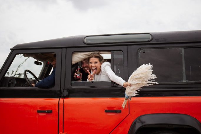 Una mujer sonriente asoma por la ventana de un coche rojo, sosteniendo un ramo.