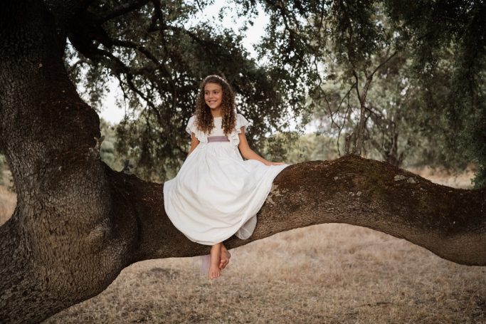 Niña sentada sobre una rama de árbol, vistiendo un vestido blanco, rodeada de naturaleza.