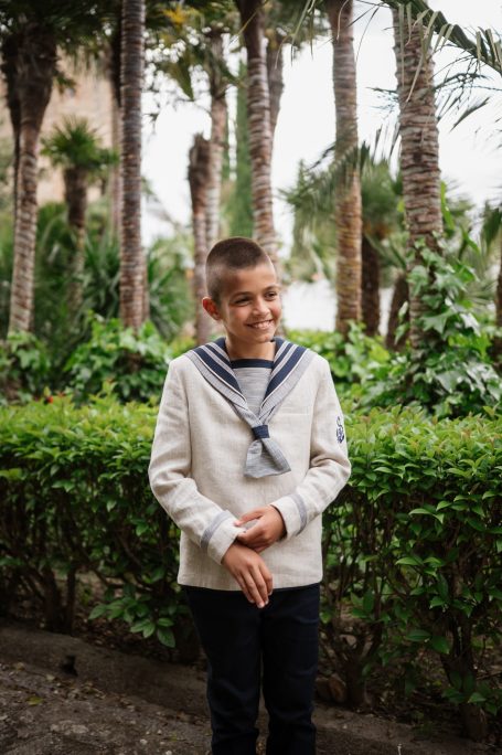 Niño sonriente con un suéter de marinero, rodeado de palmeras y vegetación.