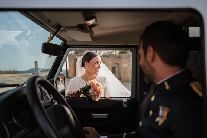 Novia sonriente con ramo de flores, mira a un hombre en un vehículo. Fondo natural.
