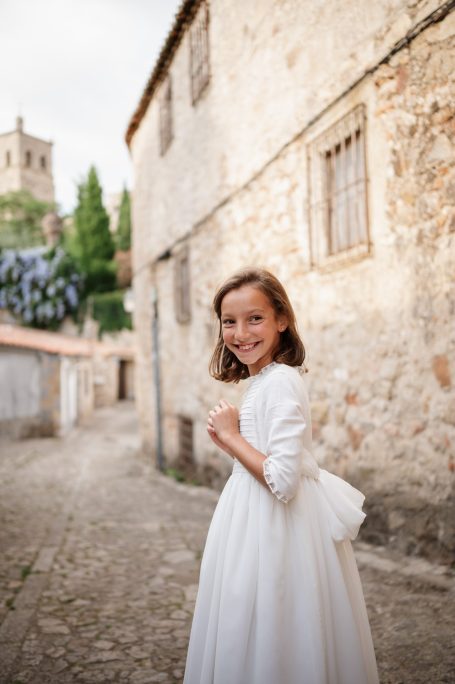 Niña con vestido blanco sonriente en un callejón de piedra, fondo de casas y vegetación.