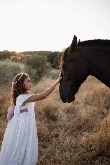 Niña en vestido blanco acariciando un caballo en un campo dorado al atardecer.